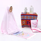 A child wearing a pink Islamic prayer dress with a blue and red patterned skirt, accompanied by a multicolored floral prayer rug, placed next to a decorative lantern and a tealight candle.