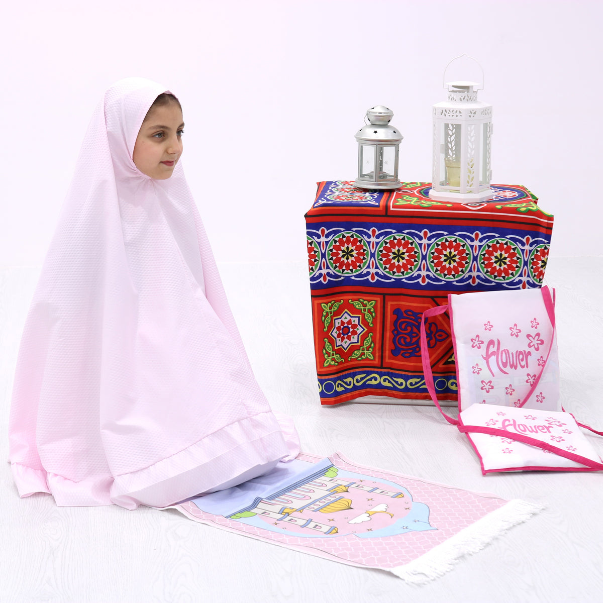 A child wearing a pink Islamic prayer dress with a blue and red patterned skirt, accompanied by a multicolored floral prayer rug, placed next to a decorative lantern and a tealight candle.